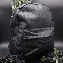 Load image into Gallery viewer, The GothX embossed skull vegan backpack on a black studio background with black roses and green foliage surrounding it. The bag is facing forward angled right to highlight the all over embossed skull design, front zip pocket and studded main zip compartment.