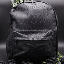 Load image into Gallery viewer, The GothX embossed skull vegan backpack on a black studio background with black roses and green foliage surrounding it. The bag is facing forward to highlight the all over embossed skull design, front zip pocket and studded main zip compartment.
