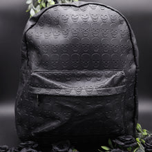 Load image into Gallery viewer, The GothX embossed skull vegan backpack on a black studio background with black roses and green foliage surrounding it. The bag is facing forward to highlight the all over embossed skull design, front zip pocket and studded main zip compartment.