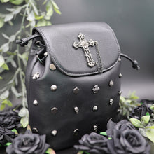 Load image into Gallery viewer, GothX dont cross me vegan tassel tie backpack on a black studio background with black roses and green foliage surrounding it. The bag is facing forward angled to the right to highlight the tassel tie cord close, hexagon, cone, cross and skull studs, and metal stud cross centrepiece with chain. 
