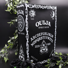 Load image into Gallery viewer, The Gothx ouija spirit book vegan backpack on a black studio background surrounded by black roses and green foliage. The bag is facing forward angled slightly right to highlight the embroidered planchette and white printed detailing featuring a ouija board, pentagrams and lace pattern.