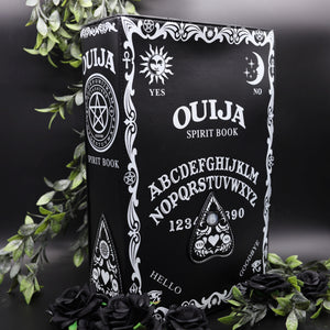 The Gothx ouija spirit book vegan backpack on a black studio background surrounded by black roses and green foliage. The bag is facing forward angled slightly right to highlight the embroidered planchette and white printed detailing featuring a ouija board, pentagrams and lace pattern.
