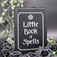 Load image into Gallery viewer, The gothx little book of spells vegan shoulder bag sat on a black studio background with green foliage and black roses surrounding it. The witchy inspired vegan leather bag is facing towards the camera to highlight the white printed design of a pentagram, framing and text reading little book of spells.