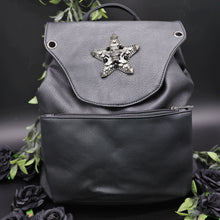 Load image into Gallery viewer, The GothX Skull and Star Black Vegan Tassel Tie Backpack in front of a black studio background with black decorative roses and leaves surrounding it. Black mini backpack bag with metal stud star with chains, faux crystals and skull head in the middle. Metal magnetic clip close and zip up large front pocket. Facing forwards to show off the detailing on the vegan leather bag.
