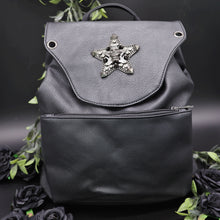 Load image into Gallery viewer, The GothX Skull and Star Black Vegan Tassel Tie Backpack in front of a black studio background with black decorative roses and leaves surrounding it. Black mini backpack bag with metal stud star with chains, faux crystals and skull head in the middle. Metal magnetic clip close and zip up large front pocket. Facing forwards to show off the detailing on the vegan leather bag.