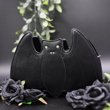 Load image into Gallery viewer, The GothX Black Bat Vegan Shoulder Bag on a black background with black roses and green foliage surrounding it. The bag is facing forward to highlight the embroidered detailing and crystal effect eyes.