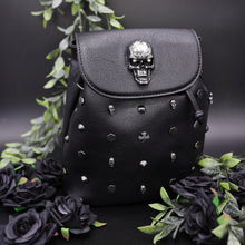 Load image into Gallery viewer, The GothX crystal skull vegan tassel tie backpack on a black studio background with black roses and green foliage surrounding it. The bag is facing forward to highlight the crystal skull magnetic close clip flap, tassel tie cords and cross, hexagon, cone and skull studs.