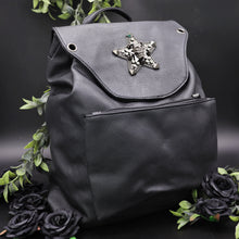 Load image into Gallery viewer, The GothX Skull and Star Black Vegan Tassel Tie Backpack in front of a black studio background with black decorative roses and leaves surrounding it. Black mini backpack bag with metal stud star with chains, faux crystals and skull head in the middle. Metal magnetic clip close and zip up large front pocket. Angled towards the right to show off the depth of the vegan leather bag.