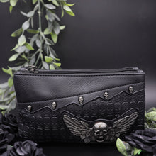 Load image into Gallery viewer, The gothx winged skull vegan clutch bag on a black studio background with black roses and green foliage surrounding it. The bag is facing forward to highlight the embossed skull leather, mini skull studs and winged skull centrepiece.