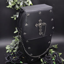 Load image into Gallery viewer, The gothx coffin vegan backpack is sat on a black studio background surrounded by black roses and green foliage. The bag is facing forward to highlight the cross mini studs, cross and chain emblem and detachable silver chain.