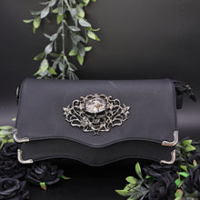 Load image into Gallery viewer, The GothX Skulls and Roses Quilted Clutch Bag sat on a black background with green foliage and black roses surrounding it. The vegan leather clutch bag is facing forward to highlight the two magnetic clip close flaps with metal corners, a stitch quilted front, a skulls and roses metal centrepiece and two D rings on either side for a detachable strap. The mini bag is inspired by gothic grunge witchy fashion.