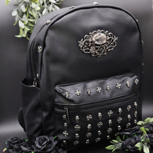 Load image into Gallery viewer, The GothX Skulls and Roses Vegan Backpack on a black studio background with black roses and foliage surrounding it. The black backpack has silver skull and cross studs along the front zip pocket and a rose skull emblem in the middle.