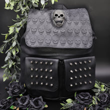 Load image into Gallery viewer, GothX twin pocket skull vegan backpack on a black background with leaves surrounding it. The bag is facing forward to highlight the diamante effect skull, skull embossed vegan leather front flap, tassel tie cords and two silver studded front pockets.
