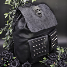 Load image into Gallery viewer, GothX twin pocket skull vegan backpack on a black background with leaves surrounding it. The bag is facing forward angled to the right to highlight the diamante effect skull, skull embossed vegan leather front flap, tassel tie cords and two silver studded front pockets.