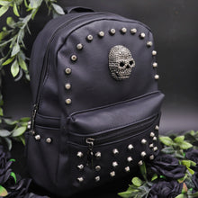 Load image into Gallery viewer, The GothX Skull Head Small Studded Vegan Mini Backpack on a black studio background with black roses and green vines surrounding them. The black vegan leather bag is facing forward angled right to highlight the diamanté style skull, gunmetal coloured studs and front zip pocket.