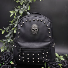 Load image into Gallery viewer, The GothX Skull Head Small Studded Vegan Mini Backpack on a black studio background with black roses and green vines surrounding them. The vegan leather bag is facing forward to highlight the diamanté style skull, gunmetal coloured studs and front zip pocket.
