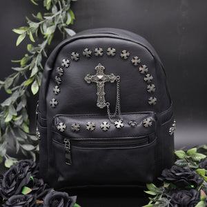 The GothX black cross vegan stud mini backpack on a black background with black roses and green leaf foliage surrounding it. The vegan leather bag is facing forwards to highlight the cross studs along the top zip line and top of zipped pocket, studded cross with chain centrepiece and side metal cross studs on the slot pockets.