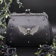 Load image into Gallery viewer, The GothX winged skull vegan vintage clasp handbag on a black studio background with black roses and green foliage surrounding it. The bag is facing forward to highlight the ball clasp close, metal floral detailing along the top, mini skull studs and winged skull centre piece.