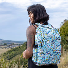 Load image into Gallery viewer, Meera is stood in a grass field wearing the kawaii unicorn vegan backpack on her back. The backpack is facing the camera, highlighting the pastel blue rainbow unicorn print, two front zip pockets, two side pockets and detachable silver chain.