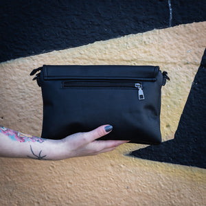 The GothX don't cross me vegan oversized clutch bag being held in front of a graffiti wall. Bag is facing away from the camera to highlight the back zip pocket and detachable strap loops.