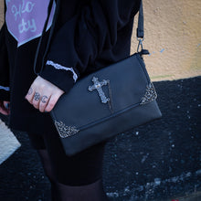 Load image into Gallery viewer, The GothX don't cross me vegan oversized clutch bag hanging on a model's shoulder with the detachable adjustable shoulder strap. Bag is facing forward to highlight the metal lace effect detailing on the front flap corners and the studded cross with chain applique.