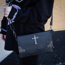 Load image into Gallery viewer, The GothX don't cross me vegan oversized clutch bag hanging on a model's shoulder with the detachable adjustable shoulder strap. Bag is facing forward to highlight the metal lace effect detailing on the front flap corners and the studded cross with chain applique.