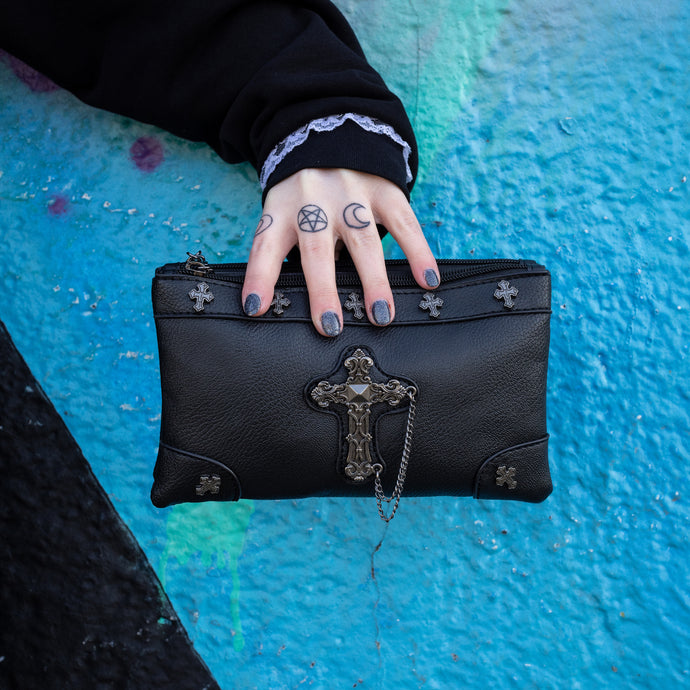 The gothx don't cross me vegan clutch bag being held by a tattooed model in front of a graffiti wall. The clutch is facing forward to highlight the cross studs and stud, chain cross centrepiece.