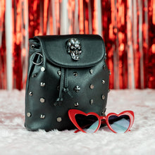 Load image into Gallery viewer, The Gothx crystal skull vegan tassel tie backpack on white fur with a red background and red heart sunglasses. The bag is facing forward to highlight the crystal skull magnetic close clip flap, tassel tie cords and cross, hexagon, cone and skull studs.
