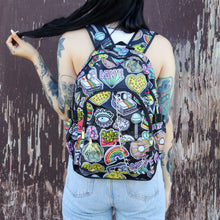 Load image into Gallery viewer, The kawaii graffiti doodle vegan backpack being worn by a tattooed model in front of a brown painted wall. The bag is facing forward to highlight the two front zip pockets, two elastic side pockets & silver chain. The all over print has lollipops, pugs, rainbows, pizzas, polaroid cameras, eyes and fries.