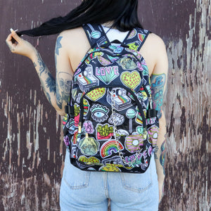 The kawaii graffiti doodle vegan backpack being worn by a tattooed model in front of a brown painted wall. The bag is facing forward to highlight the two front zip pockets, two elastic side pockets & silver chain. The all over print has lollipops, pugs, rainbows, pizzas, polaroid cameras, eyes and fries.