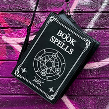 Load image into Gallery viewer, The GothX Book of Spells Vegan Messenger Bag. Vegan black leather with white witch pagan magic symbols printed on the front and spine of the 3d book bag. Bag is being held up in front of a pink and purple graffiti wall.