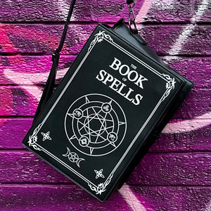 The GothX Book of Spells Vegan Messenger Bag. Vegan black leather with white witch pagan magic symbols printed on the front and spine of the 3d book bag. Bag is being held up in front of a pink and purple graffiti wall.