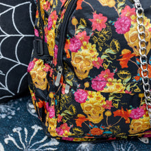 Load image into Gallery viewer, Close up of the floral gold skull nylon chain vegan backpack sat on a spooky halloween blanket next to a spider web cushion. The backpack is facing forward to highlight the gold skulls, pink and red flowers print on a black background, the two front zip pockets and two side pockets with a silver decorative chain.
