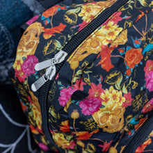 Load image into Gallery viewer, Close up of the floral gold skull nylon chain vegan backpack sat on a spooky halloween blanket next to a spider web cushion. The backpack is facing forward to highlight the gold skulls, pink and red flowers print on a black background, the two front zip pockets and two side pockets with a silver decorative chain.