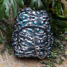 Load image into Gallery viewer, Forest Camouflage vegan backpack with chain sat outside in front of a tropical plant and brick wall. The backpack with a dark green, brown camo print is facing forward highlighting the two front zip pockets, two side pockets and silver decorative chain.