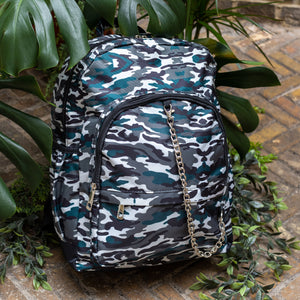 Forest Camouflage vegan backpack with chain sat outside in front of a tropical plant and brick wall. The backpack with a dark green, brown camo print is facing forward highlighting the two front zip pockets, two side pockets and silver decorative chain.