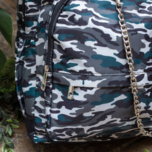 Load image into Gallery viewer, Close up of the Forest Camouflage vegan backpack with chain sat outside in front of a tropical plant and brick wall. The backpack with a dark green, brown camo print is facing forward highlighting the two front zip pockets, two side pockets and silver decorative chain.
