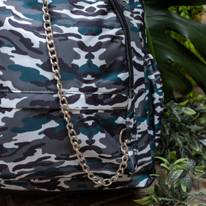 Close up of the Forest Camouflage vegan backpack with chain sat outside in front of a tropical plant and brick wall. The backpack with a dark green, brown camo print is facing forward highlighting the two front zip pockets, two side pockets and silver decorative chain.