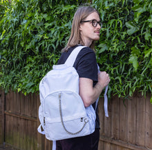 Load image into Gallery viewer, The white backpack with chain is being held by a blonde male model's shoulder whilst they look over their shoulder to the right. The vegan friendly bag is facing forward to highlight the top handle, the double zip main compartment, two front zip pockets with a silver decorative detachable draping chain and two elasticated side pockets.