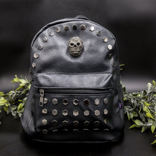 Load image into Gallery viewer, The Gothx skull head large studs vegan mini backpack on a black background with foliage surrounding it. The bag is facing forward to highlight the large hexagonal flat studded front zip pocket, diamante effect silver skull and studs along the top zip line. 
