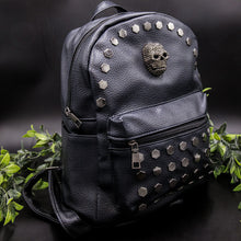 Load image into Gallery viewer, The Gothx skull head large studs vegan mini backpack on a black background with foliage surrounding it. The bag is facing forward angled right to highlight the large hexagonal flat studded front zip pocket, diamante effect silver skull and studs along the top zip line. 
