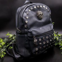 Load image into Gallery viewer, The Gothx skull head large studs vegan mini backpack on a black background with foliage surrounding it. The bag is facing forward angled right to highlight the large hexagonal flat studded front zip pocket, diamante effect silver skull and studs along the top zip line.