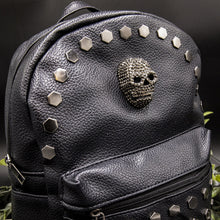 Load image into Gallery viewer, Close up of the front of the gothx skull head large studs vegan mini backpack showing the diamante effect skull and large hexagonal flat studs along the top zip line. The bag is facing forward angled to the right on a black background with foliage.