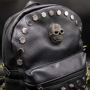 Close up of the front of the gothx skull head large studs vegan mini backpack showing the diamante effect skull and large hexagonal flat studs along the top zip line. The bag is facing forward angled to the right on a black background with foliage.