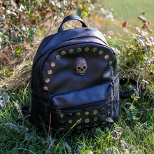 Load image into Gallery viewer, The GothX skull head large studs vegan mini backpack sat on a grass field. The bag is facing forward to highlight the large hexagonal flat studded front zip pocket, diamante effect silver skull and studs along the top zip line.