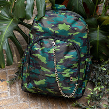 Load image into Gallery viewer, The Jungle Camouflage Backpack sat outside on a brick floor surrounded by giant monstera leaves and other green foliage. The green, brown, khaki and black vegan friendly backpack is sat facing forward to highlight the two front zip pockets with a silver draping detachable chain, the two side elasticated pockets, the top handle and the main double zip compartment.