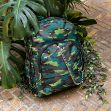 Load image into Gallery viewer, The Jungle Camouflage Backpack sat outside on a brick floor surrounded by giant monstera leaves and other green foliage. The green, brown, khaki and black vegan friendly backpack is sat facing forward to highlight the two front zip pockets with a silver draping detachable chain, the two side elasticated pockets, the top handle and the main double zip compartment.