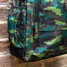 Load image into Gallery viewer, Close up of the Jungle Camouflage Backpack hanging outside on a brick wall. The green, brown, khaki and black vegan friendly backpack is facing forward to highlight the two front zip pockets with a silver draping detachable chain, the two side elasticated pockets, the top handle and the main double zip compartment.
