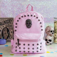 Load image into Gallery viewer, The GothX Pastel Pink Skull Head Small Studs Vegan Mini Backpack sat on an iridescent pink background with a palmistry guide poster, phrenology candle, two skulls and purple pumpkins in the background. The vegan leather bag is facing forward to highlight the dark grey metal stud detailing, diamanté effect skull, front zip pocket, two side slip pockets and top handle.