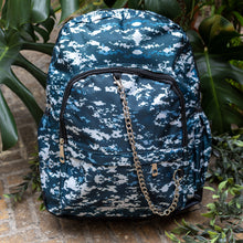 Load image into Gallery viewer, The Pixel Camouflage Backpack sat outside on a brick floor surrounded by giant monstera leaves and other green foliage. The navy, blue and white vegan friendly backpack is sat facing forward to highlight the two front zip pockets with a silver draping detachable chain, the two side elasticated pockets, the top handle and the main double zip compartment.
